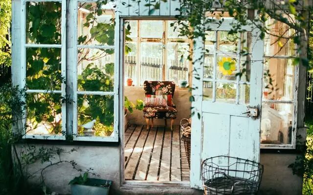 Sunlit garden shed interior with open doors, wooden floor, a patterned armchair, and greenery.