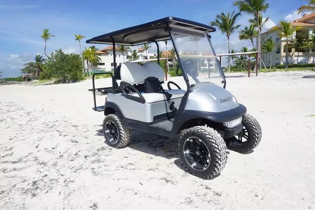 Off-road golf cart parked on a sandy beach with palm trees and resort buildings in the background.